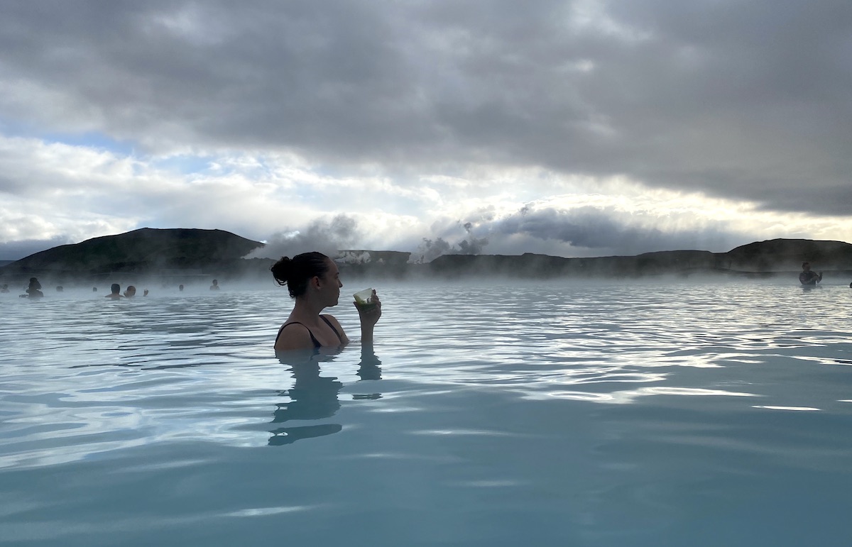 Woman drinking smoothie while soaking in bright blue lagoon overlooking lava fields on gloomy morning. Blue Lagoon, Grindavík, Iceland.