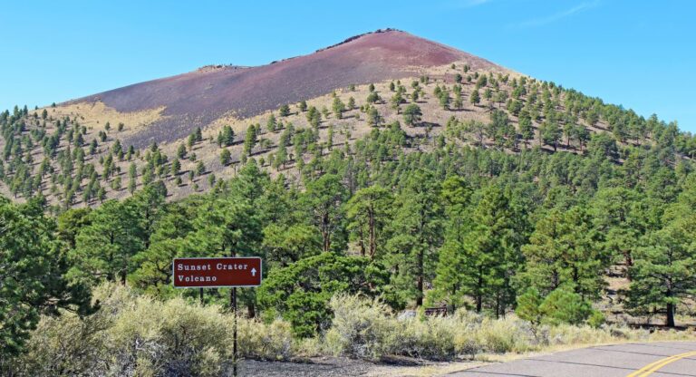 Inactive cinder volcano with red-ish tree-less top, lower portion of volcano is covered in green pine trees with road winding near volcano and sign reading "Sunset Crater Volcano" ahead. Sunset Crater Volcano National Monument, Flagstaff, Arizona.