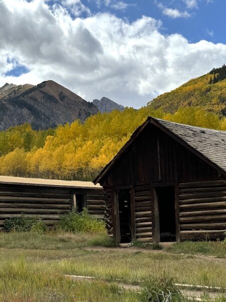 Two historic wooden buildings with yellow aspen trees and mountain peaks in background on sunny fall day. Ashcroft Ghost Down, Castle Creek Road, Aspen, Colorado.