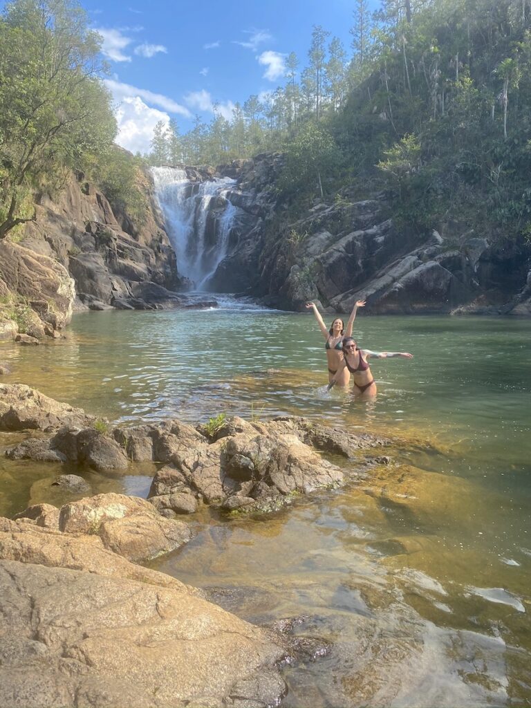 Two women smiling and posing for the camera in the water will large waterfall in background surrounded by lush green trees on sunny day. Big Rock Falls, Mountain Pine Ridge Forest Reserve, near San Ignacio, Belize.