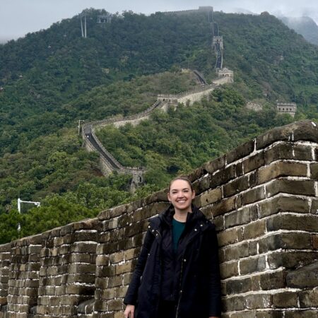 Woman leaning against wall and smiling at Great Wall of China with dramatic wall cutting through the green mountainside behind her on cloudy day. The Great Wall of China, Badaling section, Yanqing District near Beijing, China.