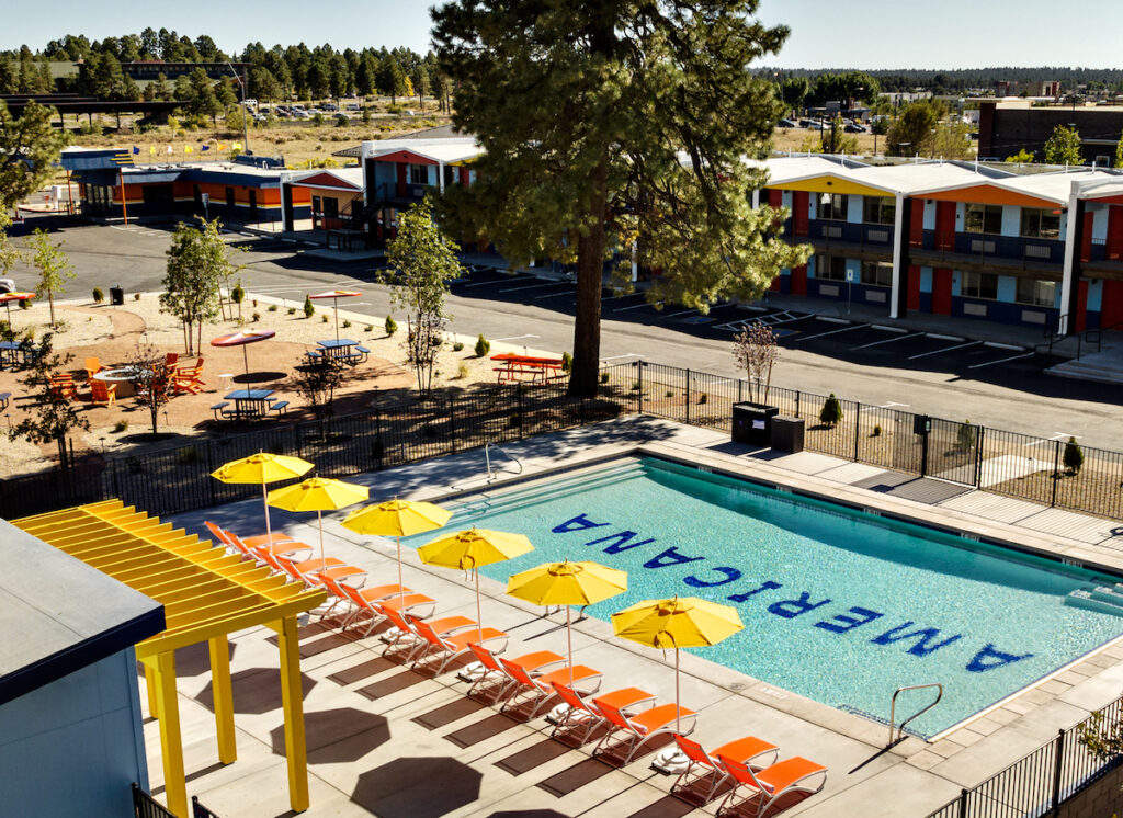 Motel pool surrounded by colorful lounge chairs and umbrellas with pool tiles reading "Americana" on sunny day. Americana Motor Hotel, Flagstaff, Arizona.