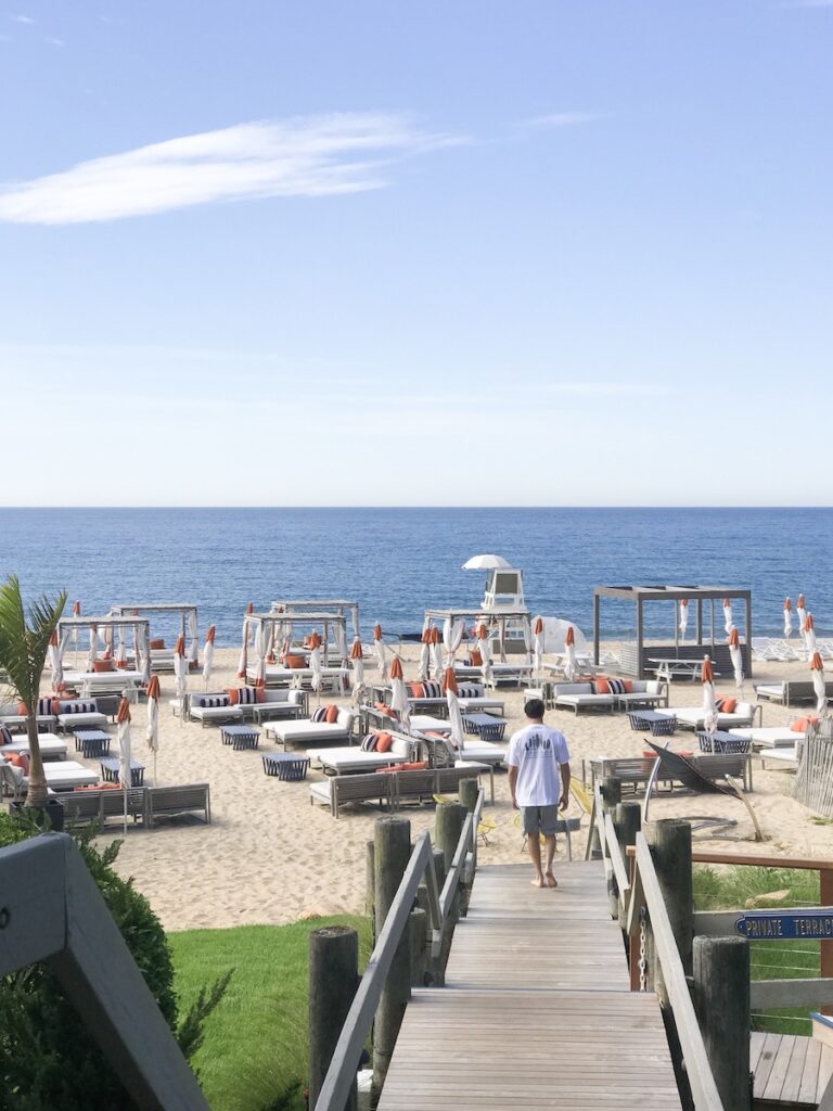 Private beach club on oceanfront beach with man walking deck leaded up to day beds and umbrellas overlooking the ocean on sunny summer day. Gurney's Montauk Resort & Seawater Spa, Montauk, New York, The Hamptons