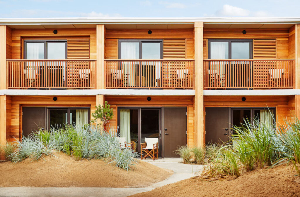 Exterior of luxury hotel with wood paneled walls, balconies, and small patios behind small sand dunes covered in American beach grass. Marram hotel, Montauk, New York, The Hamptons.