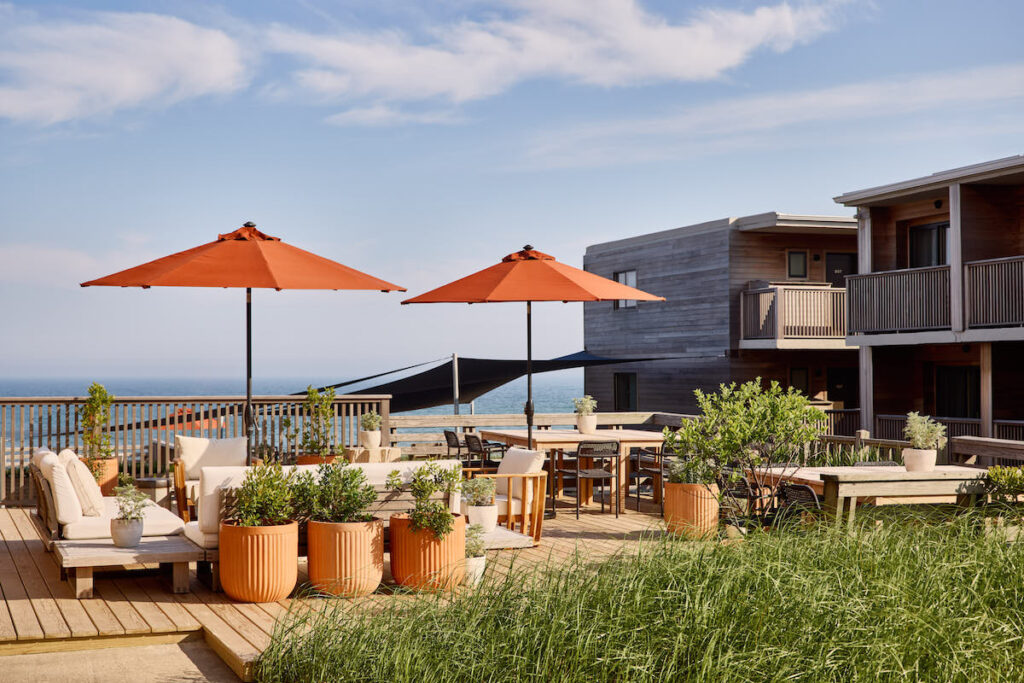 Outdoor seating area with cozy seats and umbrellas next to wood paneled luxury hotel overlooking ocean. Marram hotel, Montauk, New York, The Hamptons.