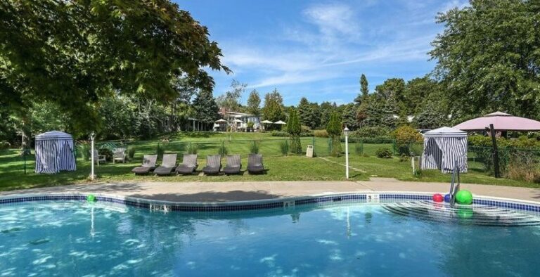 Large outdoor pool lined with lounge chairs, shaded areas, sitting on wide grassy lawn surrounded by lush green trees. Pool at Hamlet Inn, Southampton, New York, The Hamptons.