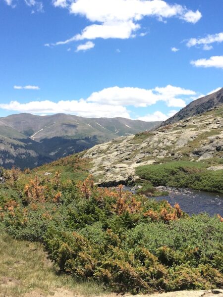 Hiking trail on sunny day with river flowing next to trail and trail opening up to expansive mountain views. McCullough Gulch, Breckenridge, Colorado.