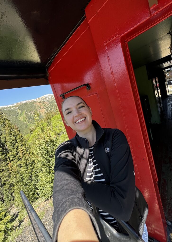 Woman smiling at camera from back of historic train car with dramatic mountain views in background on sunny summer day. Leadville Railroad, Leadville, Colorado.